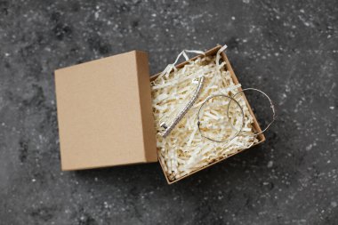 Karton kutuda gümüş küpeler ve broş var. Arka planda beton dolgu maddesi var.