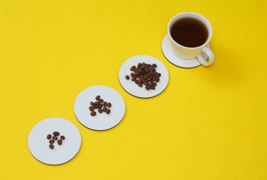 Beyaz boş bardak altlıkları ve kahve fincanı, sarı arkaplanda kahve çekirdekleri. Üst görünüm