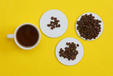 Beyaz boş bardak altlıkları ve kahve fincanı, sarı arkaplanda kahve çekirdekleri. Üst görünüm