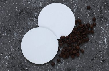 Bardaklar için beyaz boş bardak altlıkları ya da kahve çekirdekli koyu beton arka planda fincanlar.