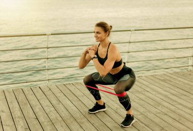 Genç bayan plaj terasında lastik bantla çömelme yapıyor.