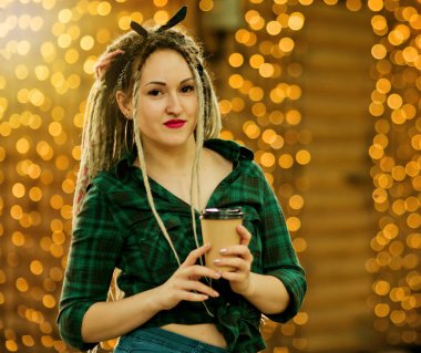 Rastgele bir saç stili ve şık yaz kıyafetleri olan gayri resmi genç bir kadın çelenklerin arka planında kahve fincanı tutuyor.