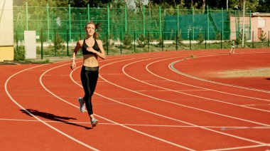 Spor kıyafetleriyle koşan neşeli genç bayan koşucu stadyum pistinde kırmızı kaplamayla koşuyor.
