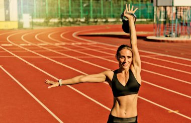 Güçlü zayıf kadın, güneşli bir günde stadyumda kırmızı kaplamalı kettlebell ile güç egzersizi yapıyor.