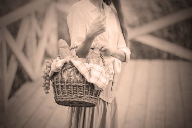 Uzun saçlı, antika elbiseli genç bir kadın gölün ahşap iskelede piknik sepeti taşıyor.