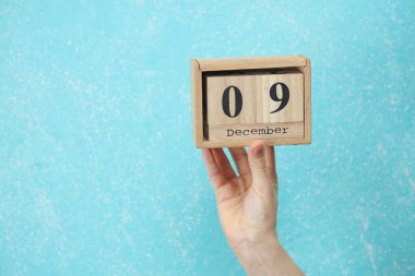 Kadının elinde ahşap blok takvimi var. Takvimde tarih 09 Aralık, mavi beton arka plan.