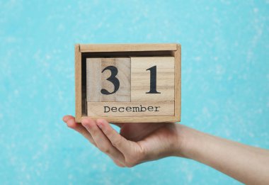 Kadının elinde ahşap blok takvimi var. Takvimin tarihi 31 Aralık, mavi beton arka plan.