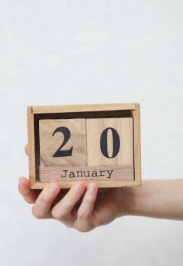 Kadının elinde tahta blok takvimi var. Tarihi 20 Ocak, beyaz beton arka planda.