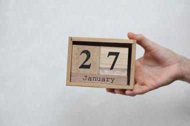 Kadının elinde tahta blok takvimi var. Tarihi 27 Ocak, beyaz beton arka planda.