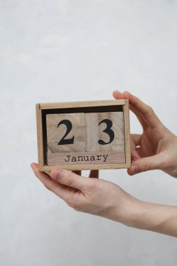 Kadının elinde tahta blok takvimi var. Tarihi 23 Ocak, beyaz beton arka planda.