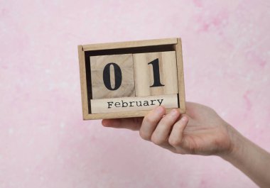 Kadının elinde tahta blok takvimi var. Tarihi 1 Şubat, pembe beton arka plan.