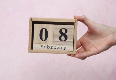 Kadının elinde ahşap blok takvimi var. Pembe beton arka planda tarih 08 Şubat.
