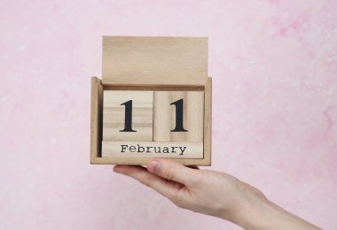 11 Şubat tarihli ahşap blok takvimi pembe beton arka planda bir kadının elinde.