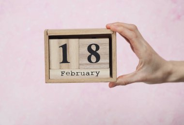 18 Şubat tarihli ahşap blok takvimi pembe beton arka planda bir kadının elinde.