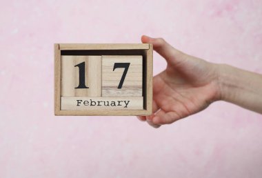 17 Şubat tarihli ahşap blok takvimi pembe beton arka planda bir kadının elinde.