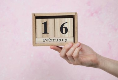 Kadının elinde tahta blok takvimi var. Tarihi 16 Şubat, pembe beton arka plan.