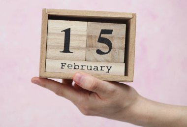 15 Şubat tarihli ahşap blok takvimi pembe beton arka planda bir kadının elinde.