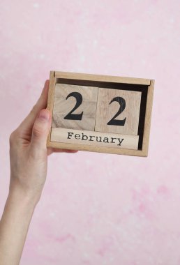 22 Şubat tarihli ahşap blok takvimi pembe beton arka planda bir kadının elinde.
