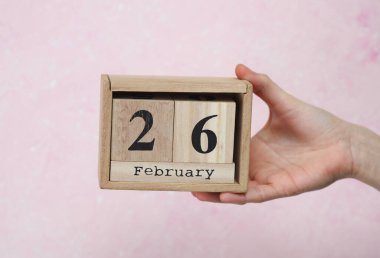 26 Şubat tarihli ahşap blok takvimi pembe beton arka planda bir kadının elinde.