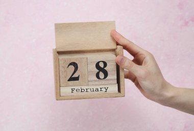 Kadının eli ahşap blok takviminde 28 Şubat tarihli pembe beton arka planda.