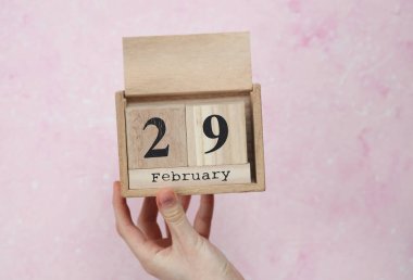 29 Şubat tarihli ahşap blok takvimi pembe beton arka planda bir kadının elinde.