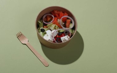 Yeşil arka planda tahta kaşıkla tek kullanımlık konteynırda Yunan salatası. Eko kavramı