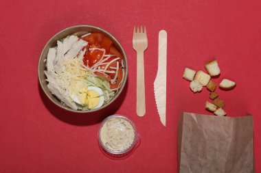 Tek kullanımlık konteynırda Sezar salatası ve kırmızı arka planda yemek aksesuarları.