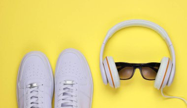 Sarı arka planda güneş gözlüklü beyaz spor ayakkabılar ve kulaklıklar. Sıradan tasarım. Müzik konsepti. Düz yatış, minimum.
