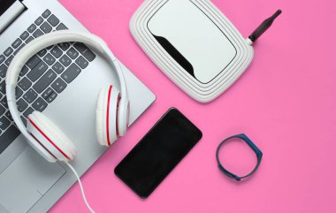 Modern dijital cihazlar ve aksesuarlar. Laptop, akıllı telefon, akıllı bilezik, kulaklık, pembe arka planda kablosuz ağ yönlendirici. Üst görünüm.