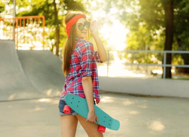 Parlak güneşli bir günde kaykay parkında elinde kaykay tutarken şık giyinmiş neşeli, duygusal genç bir kadın.