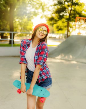 Parlak güneşli bir günde kaykay parkında elinde kaykay tutarken şık kıyafetler giymiş neşeli genç bir kadın.