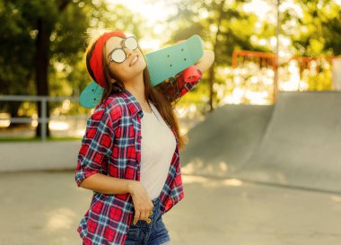 Parlak güneşli bir günde kaykay parkında elinde kaykay tutarken şık giyinmiş neşeli, duygusal genç bir kadın.