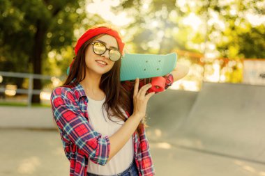 Parlak güneşli bir günde kaykay elinde kaykay tutarken şık kıyafetler ve aynalı güneş gözlüğü takan neşeli, duygusal genç bir kadın.