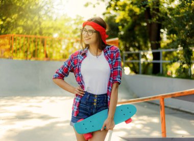 Parlak güneşli bir günde kaykay parkında şık kıyafetler giymiş neşeli genç hippi kadın elinde kaykayla geziyor.