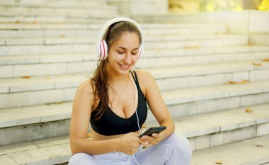Eğitimden sonra dinlenme kavramı. Spor giyim sektöründe genç ve mutlu bir kadın akıllı telefon kullanıyor ve güneşli bir günde merdivenlerde otururken kulaklıkla müzik dinliyor.