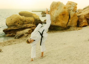 Siyah kuşaklı trenleri olan beyaz kimonolu genç, çekici bir karateci kadın taşlarla dolu vahşi bir plaja tekme atıyor.