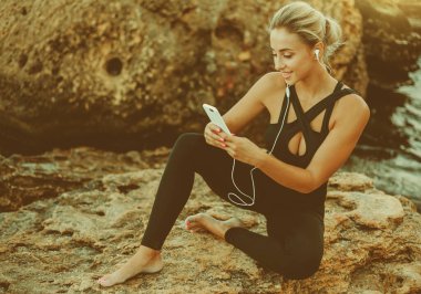 Spor elbiseli genç bir kadın kulaklıkla müzik dinliyor ve vahşi sahilde bir taşın üzerinde otururken akıllı telefon kullanıyor.