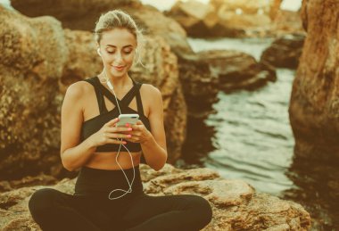 Spor elbiseli genç bir kadın kulaklıkla müzik dinliyor ve vahşi sahilde bir taşın üzerinde otururken akıllı telefon kullanıyor.