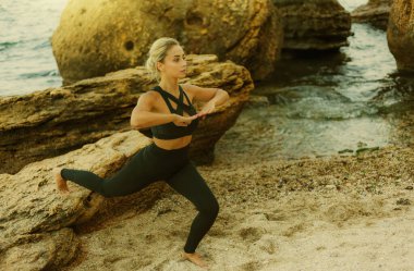 Spor kıyafetli genç, zayıf bir kadın vahşi sahilde taş üzerinde jimnastik yapıyor. Isının..