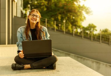 Merdivenlerde dizüstü bilgisayarla oturan kot ceketli, modern bir bayan öğrenci. Mesafe öğrenme. Modern gençlik kavramı.
