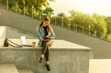Genç çekici öğrenci kadın kentteki merdivenlerde otururken bir kitap okuyor. Mesafe öğrenme kavramı. Modern öğrenci