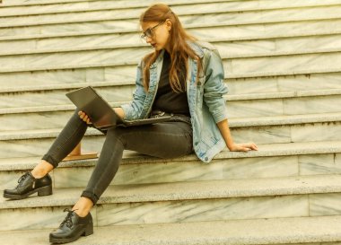 Merdivenlerde dizüstü bilgisayarla oturan kot ceketli, modern bir bayan öğrenci. Video izliyorum. Mesafe öğrenme. Modern gençlik kavramı.