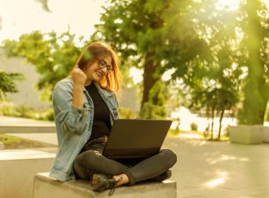 Kot ceketli genç ve mutlu bayan öğrenci parkta oturuyor ve dizüstü bilgisayarın ekranına bakıyor. Mesafe öğrenme. Çevrimiçi çağrı. Modern gençlik kavramı.