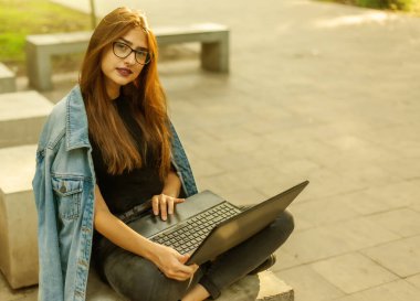 Merdivenlerde dizüstü bilgisayarla oturan kot ceketli, modern bir bayan öğrenci. Mesafe öğrenme. Modern gençlik kavramı.