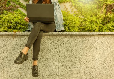 Kot ceketli ve gözlüklü mutlu bayan öğrenci şehir parkında otururken dizüstü bilgisayar kullanıyor. Kırpılmış fotoğraf