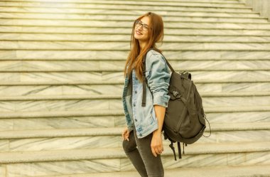 Kot ceketli, gözlüklü, büyük sırt çantalı neşeli kız öğrenci şehirde merdivenlerden çıkıyor. Gençlik kavramı