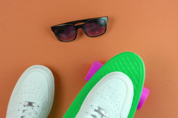 White sneakers on plastic mini cruiser board and sunglasses. Brown background. Summertime fun. Top view