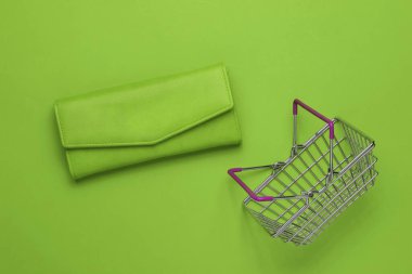 Minimalist alışveriş konsepti. Mini alışveriş sepeti ve yeşil arka planda yeşil deri cüzdan. Üst görünüm