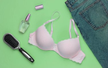 Mavi kot, sutyen, parfüm şişesi, yeşil arka planda tarak. Kadının günlük kıyafetleri ve aksesuarları. Moda kadın kıyafetleri. Düz yatış, üst görünüm.