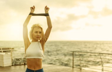 Deniz kenarındaki güvertede sağlıklı kadın eğitimi. Spor giyimli bir kadın güneş doğarken plajda spor lastiğiyle egzersiz yapıyor.
